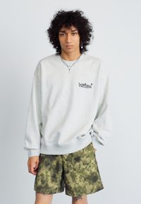 Sudadera gris claro con puños y dobladillo acanalados, que presenta un pequeño logotipo negro. Un par de pantalones cortos de camuflaje verde con un ajuste relajado.