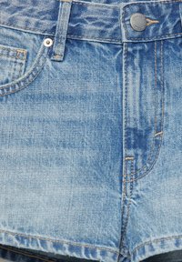 Gros plan d'un short en denim bleu clair montrant le bouton avant, la fermeture éclair, la boucle de ceinture et les poches avant avec des détails de couture visibles.