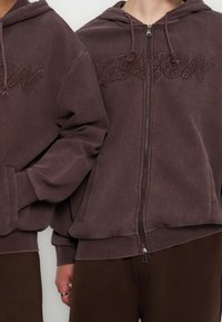 Δύο άτομα φορούν καφέ υφασμάτινα φούτερ με φερμουάρ και κεντημένο το «ASTON» στο στήθος, συνδυασμένα με καφέ παντελόνια.