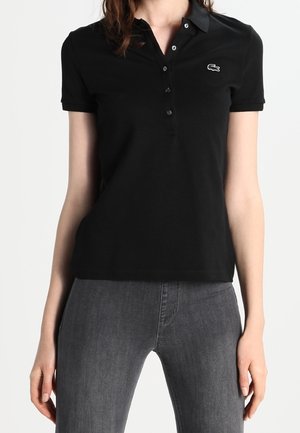 Kvinde iført en sort kortærmet poloshirt med knapper og et lille hvidt krokodille-logo, kombineret med mørkegrå jeans.