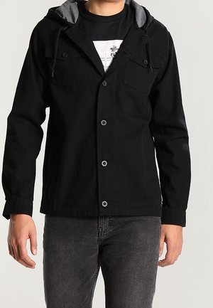 Mand iført en sort knappet jakke med fasthætte hætte over en sort grafisk T-shirt og mørkegrå jeans, stående mod en ensfarvet baggrund.