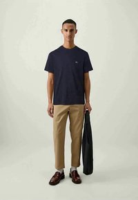 Tricou bleumarin cu mânecă scurtă, guler rotund și un logo mic, combinat cu pantaloni bej adaptați și pantofi negri, purtând un sacou.