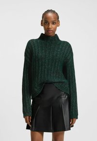 Grön stickad tröja med hög krage, ribbad textur och lös passform, parat med en svart veckad läderskjorta.