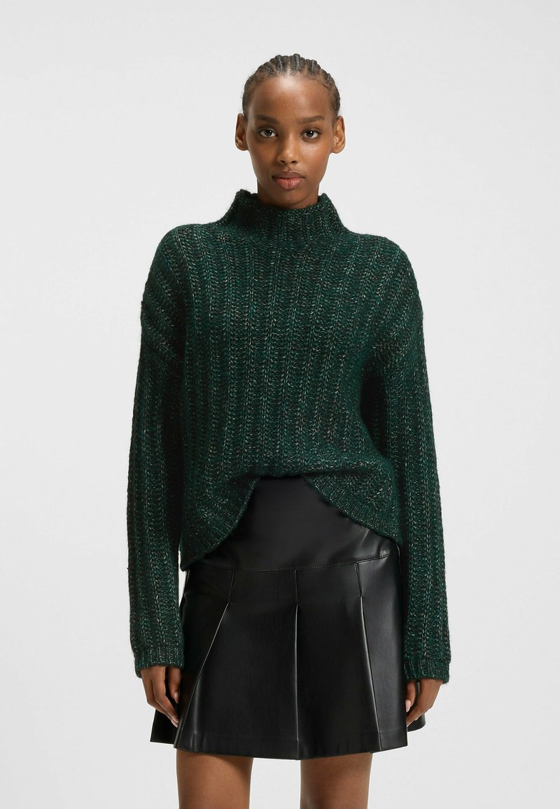 Grön stickad tröja med hög krage, ribbad textur och lös passform, parat med en svart veckad läderskjorta.