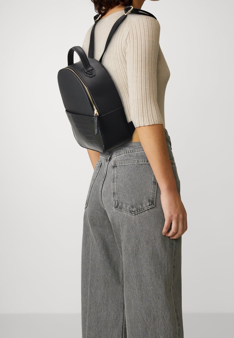 Femme portant un haut beige côtelé et un jean gris, portant un petit sac à dos en cuir noir avec une poche avant zippée.
