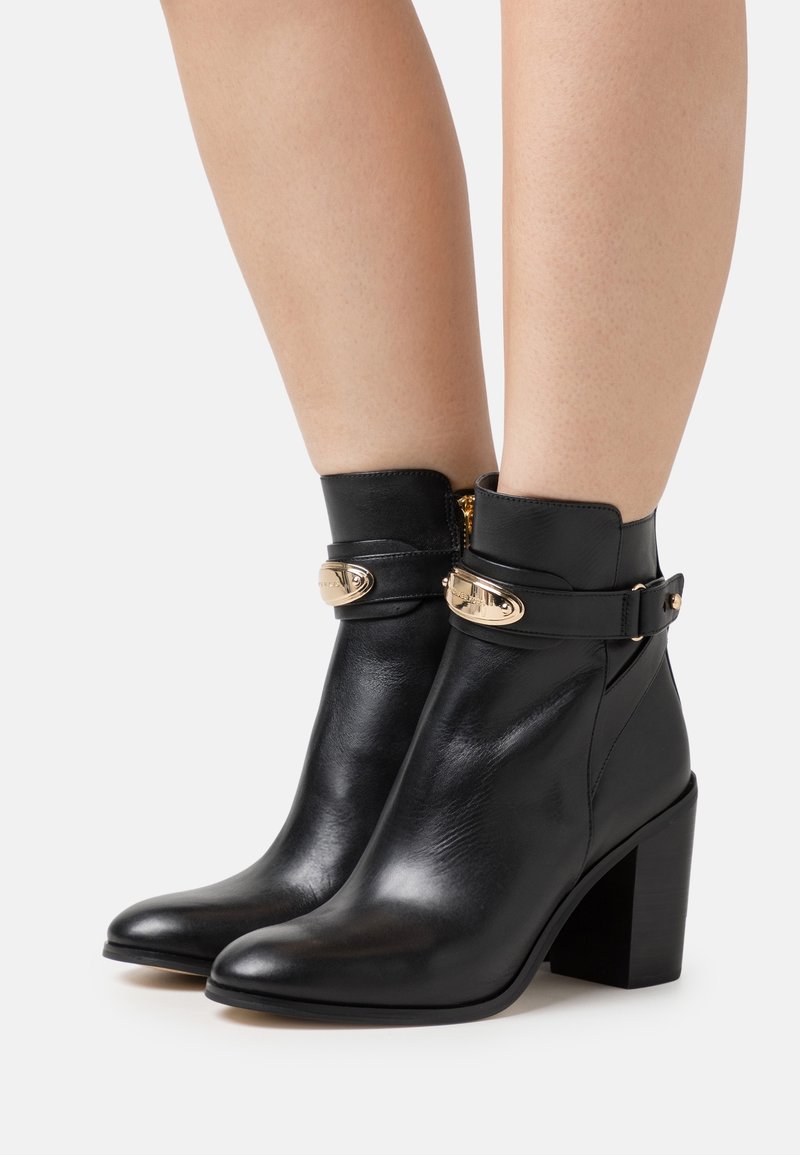 Bottes courtes en cuir noir avec talons blocs et détails de boucles dorées portées sur des jambes nues sur un fond clair uni.