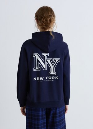 Person mit braunen Haaren im Dutt trägt einen dunkelblauen Hoodie mit großem weißen Text "NY" und "New York" auf dem Rücken, kombiniert mit blau karierten Hosen.