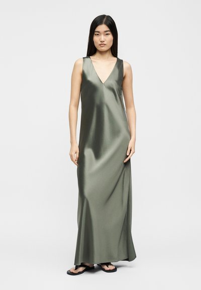 Tânără cu păr lung și negru, purtând o rochie maxi din satin verde măsliniu fără mâneci și sandale negre, stând în picioare pe un fundal simplu, deschis la culoare.