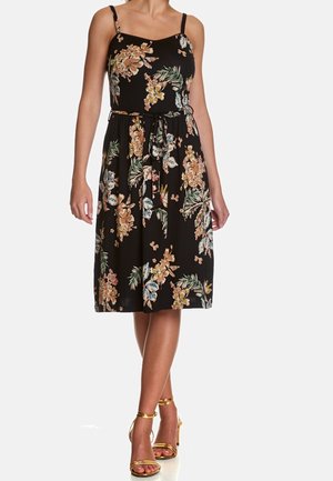 Femme portant une robe midi noire à fleurs sans manches avec une ceinture à la taille et des talons dorés à brides, debout, jambes croisées.