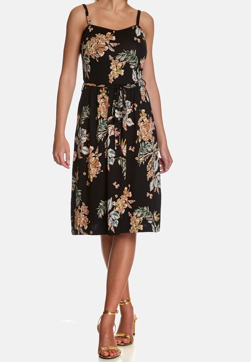 Femme portant une robe midi noire à fleurs sans manches avec une ceinture à la taille et des talons dorés à brides, debout, jambes croisées.