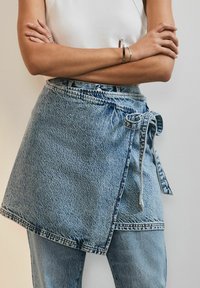 Sirena od jeansa u svijetloplavoj boji, s detaljem vezivanja u struku. Izrađena od teksturiranog jeansa s izfrayanim rubom i preklopnim dizajnom.
