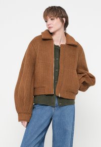 Brun cropped jakke med tekstureret stof, krave og ribbet kant. Båret over en grøn knappet top og parret med blå jeans.