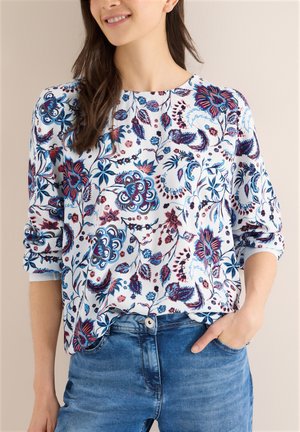 Kvinde iført bluse med blå og rødt blomsterprint, langærmet, stoppet ned i højtaljede blå jeans med den ene hånd i lommen.