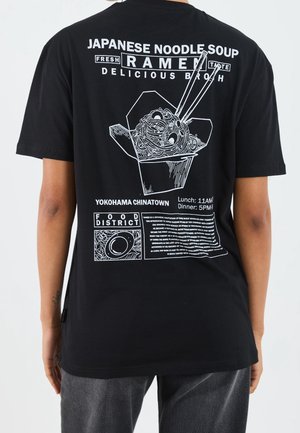 T-shirt en coton noir avec un imprimé graphique blanc représentant des nouilles ramen dans une boîte à emporter, avec du texte et des illustrations détaillées au dos.