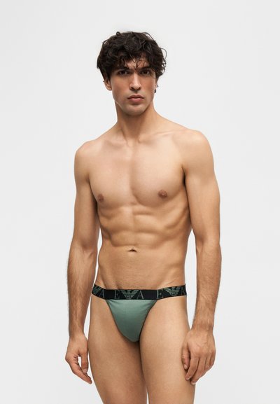 Jeune homme musclé portant un sous-vêtement vert avec une ceinture noire comportant un logo, debout devant un fond clair uni.