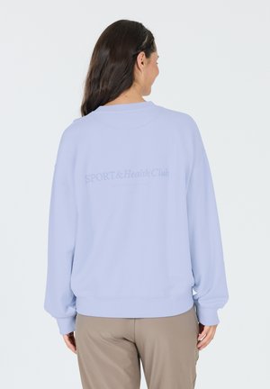 Sudadera de gran tamaño en azul claro con puños y dobladillo acanalados. Texto "SPORT & Health Club" impreso en un tono más claro en la parte posterior. Textura de tela suave.