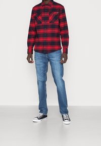 Camicia di flanella a quadri rossi e blu con due tasche sul petto, abbinata a jeans blu e sneakers alte nere. Texture morbide e vestibilità casual.