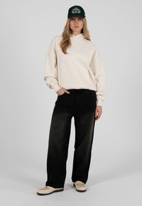 Felpa con cappuccio beige con tasca a marsupio, jeans neri a gamba larga e scarpe da ginnastica color crema. Cappellino verde con testo ricamato. Outfit semplice e casual.