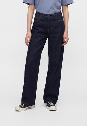 Persona che indossa jeans a gamba larga blu scuro, scarpe da ginnastica nere e bianche e una camicia a maniche corte azzurro chiaro, in piedi davanti a uno sfondo bianco.