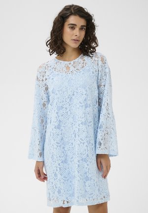 Kobieta o kręconych włosach, ubrana w jasnoniebieską, kwiecistą sukienkę z koronki z długim rękawem, stojąca na gładkim białym tle.