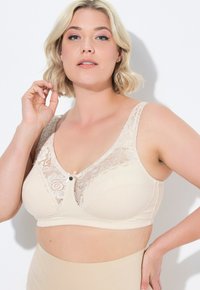 Beige beha met kantdetails aan de bovenkant, met een bloemenpatroon en een centrale decoratieve knop. Beschikt over brede banden en een zachte stoftextuur.