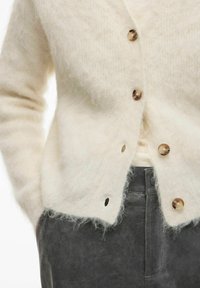 Kremasta pletena jopica z mehkim zaključkom, z velikimi svetlo rjavimi gumbi in stranskimi žepi, nošena preko temno sivih hlač.