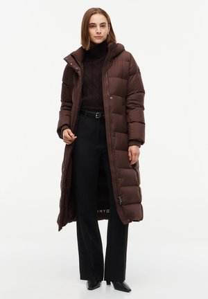 Femme portant un long manteau matelassé marron par-dessus un col roulé noir et un pantalon noir, debout sur un fond blanc.
