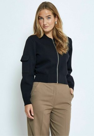 Black cropped zip-up cardigan with ribbed hem and long sleeves, featuring a side pocket on the left sleeve. Worn with beige trousers.