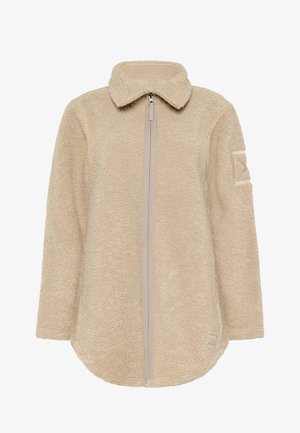 Beżowa kurtka polarowa na całym zamek z wysokim kołnierzem, długimi rękawami, teksturowaną tkaniną i małą naszywką na lewym górnym ramieniu.