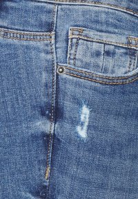 Detailní záběr modrých džín ukazující švy, nýtek, malou přední kapsu a opotřebovanou, poškrábanou náplast blízko švu.