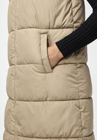 Beige vadderad väst med en sidoficka, horisontell sömnad och en ribbad svart tröjärm delvis synlig. Mjuk textur.