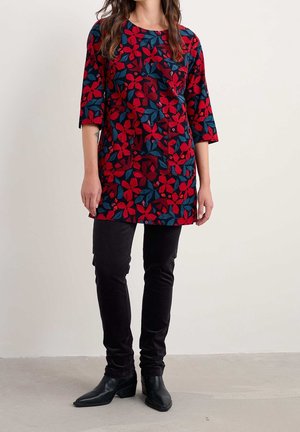 Kobieta ubrana w czerwoną i granatową kwiatową tunikę z rękawami do połowy przedramienia, czarne dopasowane spodnie oraz czarne botki, stojąca na szarej podłodze.