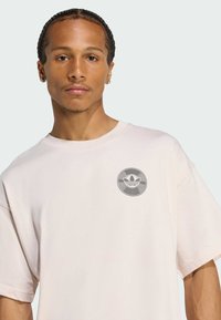 Ljusrosa bomullst-shirt med korta ärmar, med en cirkulär grå logotyp på bröstet. Mjuk textur och avslappnad passform.