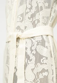 Κοντινό πλάνο λευκής διάφανης δαντέλας με λουλουδάτα μοτίβα και μια ζώνη από το ίδιο ύφασμα δεμένη σε κόμπο στη μέση.