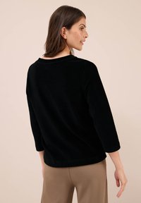 Vrouw die zijwaarts kijkt, gekleed in een zwart langemouwen top en een beige broek, tegen een effen beige achtergrond.