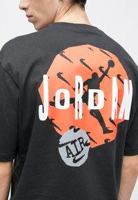 Μαύρο βαμβακερό t-shirt με μεγάλο πορτοκαλί εξάγωνο γραφικό που περιλαμβάνει τη σιλουέτα ενός μπάσκετ, τα λόγια "JORDAN" και "AIR" σε λευκό και γκρι.