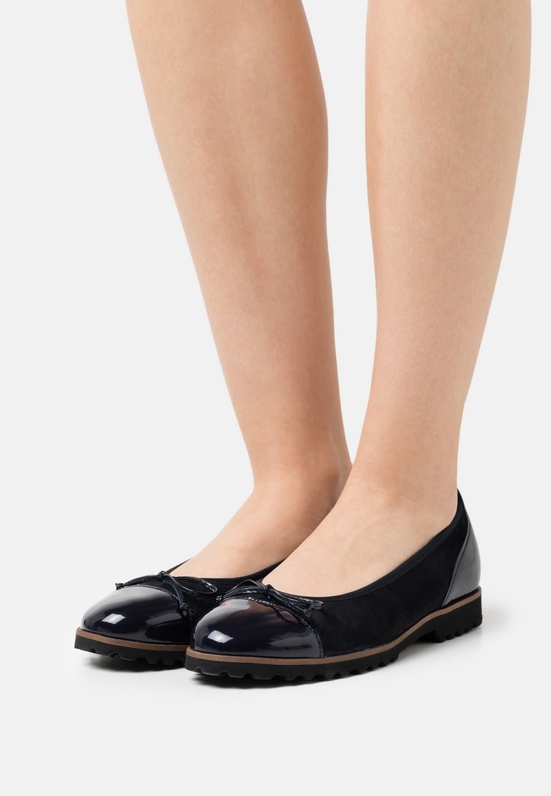 Paire de jambes portant des ballerines noires avec des bouts brillants et de petits détails en forme de nœud sur un fond blanc.