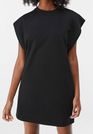 Kobieta w czarnej mini sukience bez rękawów z poduszkami na ramionach, stojąca na tle jednolitego białego tła.