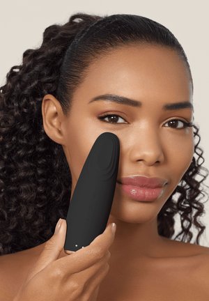 Woman with curly hair holds a black facial cleansing device to her right cheek, demonstrating skincare application.