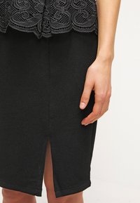 Femme portant une jupe crayon texturée noire avec une fente à l'avant et un haut en dentelle noire, la main reposant le long du corps.