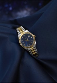 Sølv- og gullfarget klokke med en marineblå urskive, rustfritt stålkasse og armbånd med et strukturert mønster med lenker.