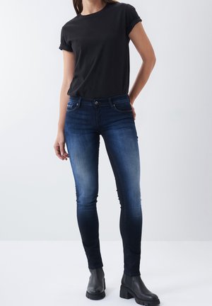 Kobieta ubrana w czarny t-shirt z podwiniętymi rękawami, ciemnoniebieskie obcisłe dżinsy i czarne botki, stojąca z jedną ręką za plecami.