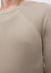 Pull en maille côtelée beige avec des détails de côtes diagonales sur l'épaule et un col rond porté par une personne.
