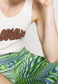 Bílý tílko s hnědým nápisem "OUTSIDE", doplněné zelenými a modrými kalhotami se vzorem vlnitých čar. Materiál se jeví jako hladký a přiléhavý.