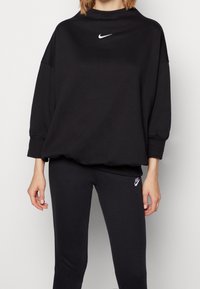 Svart sweatshirt med hög krage och vitt Nike-logga, i kombination med svarta leggings som har en liten Nike-logga på benet. Mjuk tyg.