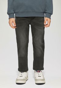 Enfant portant un jean noir délavé, des baskets blanches avec des détails rouges et bleus, et un haut à manches longues gris foncé, se tenant devant un fond uni.