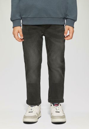 Kind gekleed in vervaagde zwarte jeans, witte sneakers met rode en blauwe details, en een donkergrijze lange mouwtop, staande tegen een effen achtergrond.