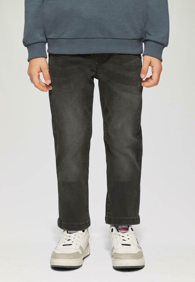 Enfant portant un jean noir délavé, des baskets blanches avec des détails rouges et bleus, et un haut à manches longues gris foncé, se tenant devant un fond uni.