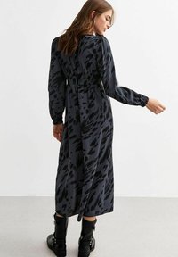 Langes, schwarzes Kleid mit Leopardenmuster, ausgestattet mit langen Ärmeln und tailliertem Bund. Hergestellt aus glattem Stoff, kombiniert mit schwarzen Knöchelstiefeln.