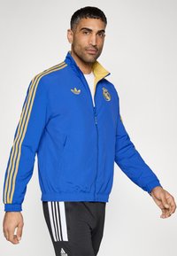 Blue zip-up jacket with gold accents, featuring a Real Madrid logo and three gold stripes on sleeves. Made of lightweight fabric, elastic cuffs.